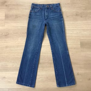 May include: A pair of blue denim jeans with a classic five-pocket design. The jeans have a button and zipper closure, belt loops, and a straight leg fit. The denim has a faded appearance, suggesting wear. The jeans are laid flat on a light-colored wooden surface.