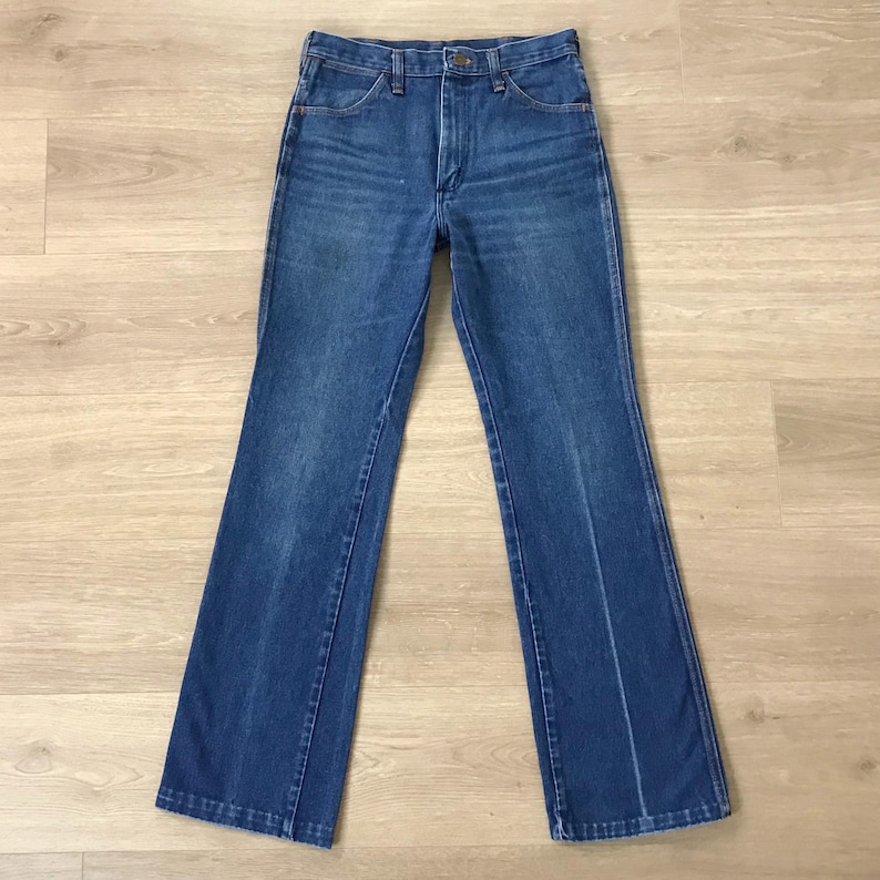 May include: A pair of blue denim jeans with a classic five-pocket design. The jeans have a button and zipper closure, belt loops, and a straight leg fit. The denim has a faded appearance, suggesting wear. The jeans are laid flat on a light-colored wooden surface.