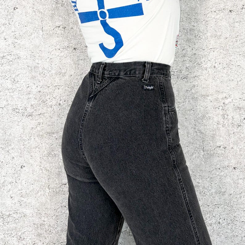 Pu&ograve; includere: Primo piano di jeans neri. I jeans hanno una vita alta e un classico design a cinque tasche. Il logo Wrangler &egrave; visibile sulla tasca posteriore. I jeans sono indossati con una maglietta bianca con una grafica blu.
