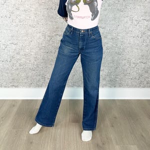 May include: Dark wash denim jeans with a classic five-pocket design. The jeans have a straight leg fit and a button and zipper closure. The model is wearing a white t-shirt with a graphic print and white socks.