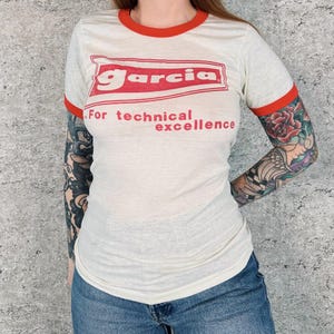 May include: Cream ringer t-shirt with red trim and text. The shirt features the word "Garcia" in a red rectangular box above the phrase "For technical excellence" in red. The shirt is worn with blue jeans.