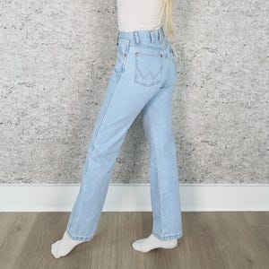 May include: Pale blue denim jeans with a classic five-pocket design. The jeans have a high waist and a straight leg fit. The back pocket features a stitched logo. The jeans are worn with a white top and white socks.