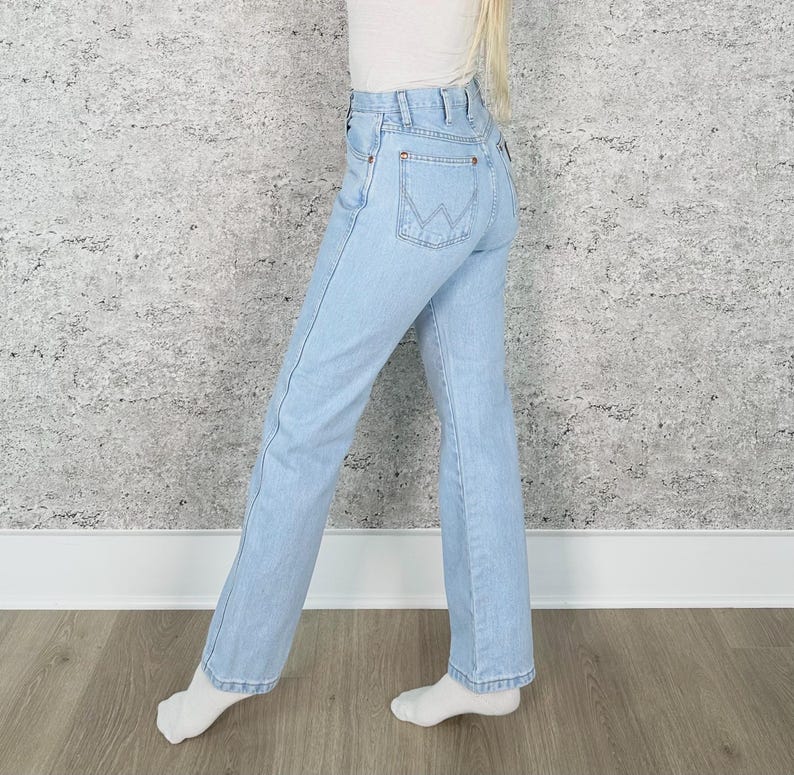 May include: Pale blue denim jeans with a classic five-pocket design. The jeans have a high waist and a straight leg fit. The back pocket features a stitched logo. The jeans are worn with a white top and white socks.