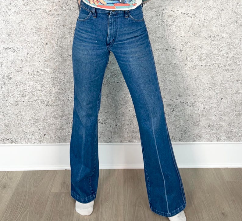 May include: Blue denim bell-bottom jeans. The jeans have a classic five-pocket design and a high waist. The legs flare out from the knee to the hem. The jeans are a medium wash and have a slight fade.