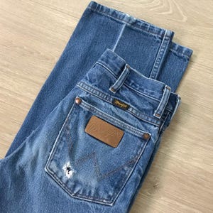 May include: Blue denim jeans featuring a brown leather patch on the back pocket with the Wrangler logo. The jeans include copper-coloured rivets, belt loops, and a distinctive W-shaped stitching pattern on the pocket, along with a small distressed hole.
