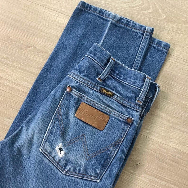 May include: Blue denim jeans featuring a brown leather patch on the back pocket with the Wrangler logo. The jeans include copper-coloured rivets, belt loops, and a distinctive W-shaped stitching pattern on the pocket, along with a small distressed hole.