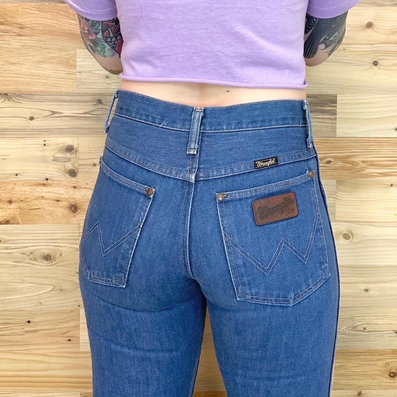Pu&ograve; includere: Jeans blu in denim con vita alta e toppa Wrangler sulla tasca posteriore. I jeans hanno un taglio dritto e un'estetica vintage. I jeans sono di un blu medio.