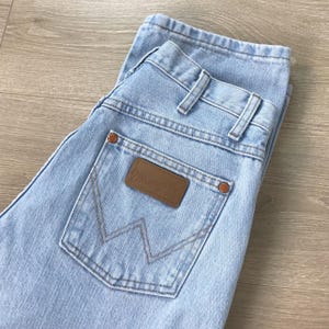 May include: Pale blue denim jeans featuring a brown leather patch on the back pocket. The jeans have a classic five-pocket design and a straight leg fit. The brand name "Wrangler" is visible on the patch.