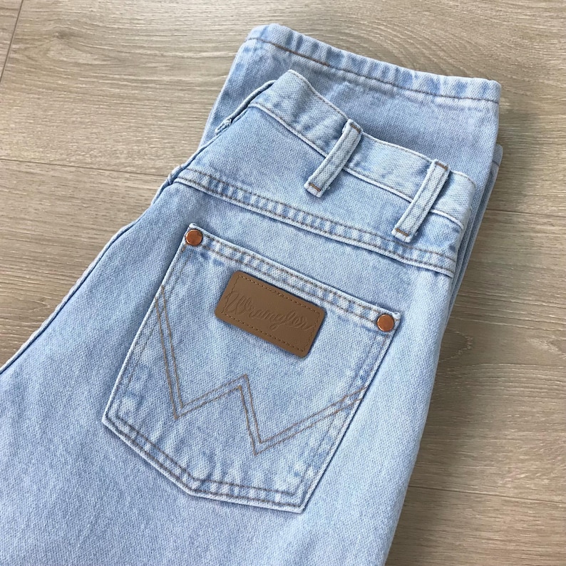 May include: Pale blue denim jeans featuring a brown leather patch on the back pocket. The jeans have a classic five-pocket design and a straight leg fit. The brand name "Wrangler" is visible on the patch.