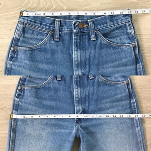 Op de afbeelding: Close-up van lichtblauwe denim jeans met een knoopsluiting en zakken. Een meetlint is over de taille geplaatst, met een meting van ongeveer 35.5 cm. De jeans heeft een klassieke, vintage stijl.