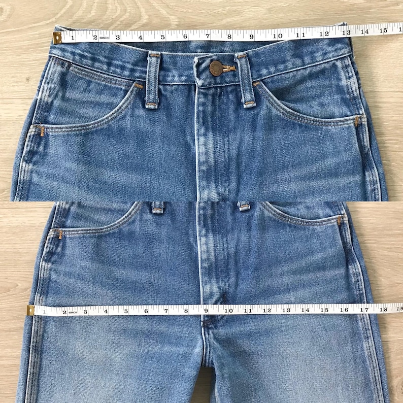 Op de afbeelding: Close-up van lichtblauwe denim jeans met een knoopsluiting en zakken. Een meetlint is over de taille geplaatst, met een meting van ongeveer 35.5 cm. De jeans heeft een klassieke, vintage stijl.