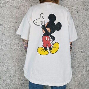 Op de afbeelding: Wit T-shirt met een grote Mickey Mouse-afbeelding. Het stripfiguur is afgebeeld in zwart, rood, geel en wit, zwaaiend. Het shirt heeft korte mouwen en een losse pasvorm. Het shirt is gemaakt van een zacht materiaal.