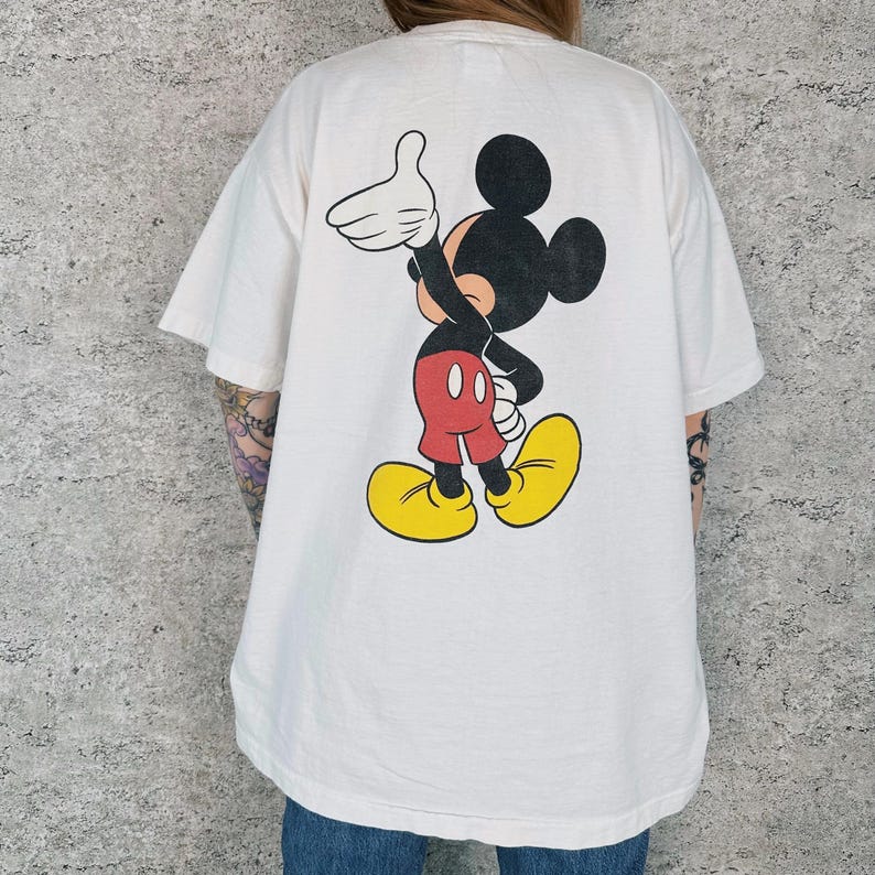 Op de afbeelding: Wit T-shirt met een grote Mickey Mouse-afbeelding. Het stripfiguur is afgebeeld in zwart, rood, geel en wit, zwaaiend. Het shirt heeft korte mouwen en een losse pasvorm. Het shirt is gemaakt van een zacht materiaal.