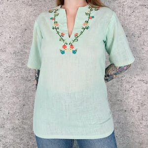 May include: A light mint green short-sleeved shirt featuring a V-neckline and intricate floral embroidery in shades of red, orange, yellow, and blue along the collar. The garment has a textured fabric finish and a straight hemline.
