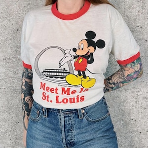May include: White ringer t-shirt with red trim featuring a graphic of Mickey Mouse, the Gateway Arch, and the text "Meet Me in St. Louis." The shirt has short sleeves and a classic fit.