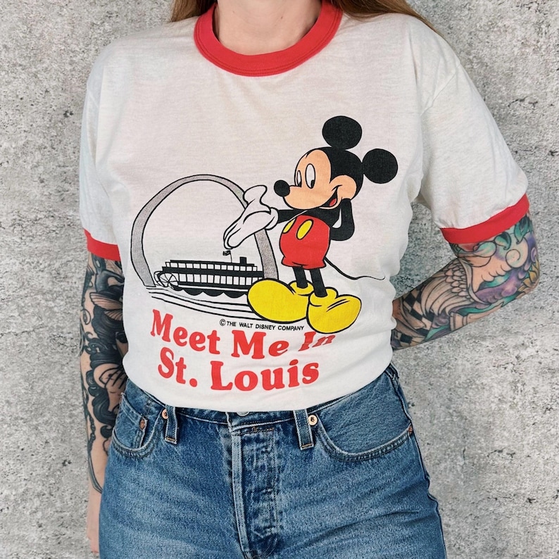 May include: White ringer t-shirt with red trim featuring a graphic of Mickey Mouse, the Gateway Arch, and the text "Meet Me in St. Louis." The shirt has short sleeves and a classic fit.