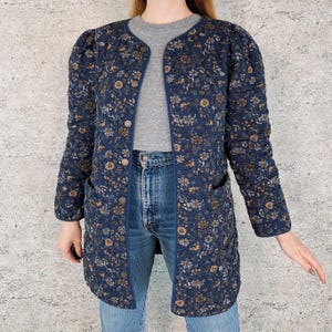 May include: A navy blue quilted jacket with a floral pattern. The jacket has a round collar, long sleeves, and a button-up front. It features two front pockets and is approximately 40 inches long.