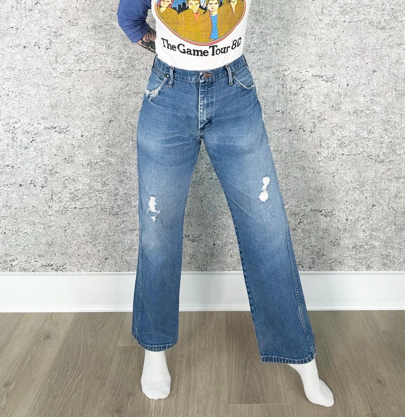 K&ouml;nnte beinhalten: Blaue Jeans aus Denim mit Distressed-Details. Die Jeans haben eine hohe Taille und ein weites Bein. Das Model tr&auml;gt ein wei&szlig;es und gelbes Grafik-T-Shirt mit dem Text "The Game Tour 80".