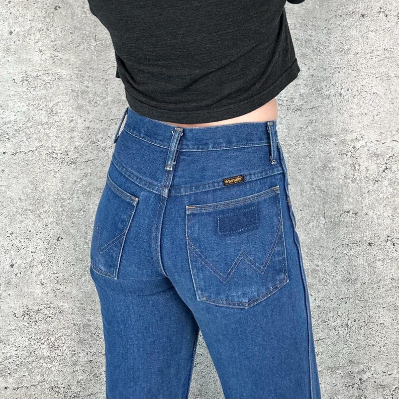 May include: A pair of blue denim jeans with a high waist and a classic straight leg fit. The jeans have a back pocket with a stitched-on Wrangler logo.