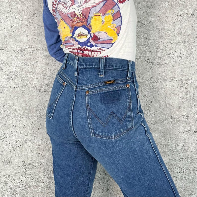 K&ouml;nnte beinhalten: Blaue Wrangler Jeans mit hohem Bund und klassischem R&uuml;ckentaschendesign. Die Jeans haben einen geraden Beinverlauf und das Wrangler-Logo auf der Ges&auml;&szlig;tasche. Das Model tr&auml;gt ein lang&auml;rmliges Shirt mit einem grafischen Design.