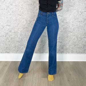 May include: Dark blue denim jeans with a high waist and flared leg. The jeans have a classic five-pocket design and a button and zipper closure. The model is wearing a black top and yellow socks.