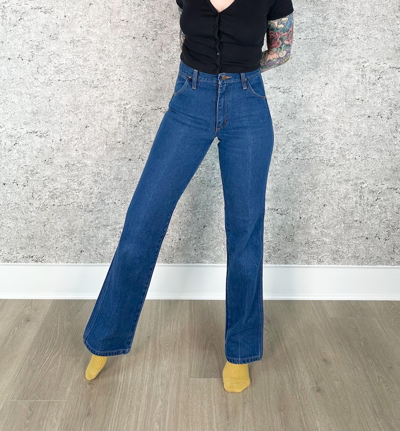 May include: Dark blue denim jeans with a high waist and flared leg. The jeans have a classic five-pocket design and a button and zipper closure. The model is wearing a black top and yellow socks.