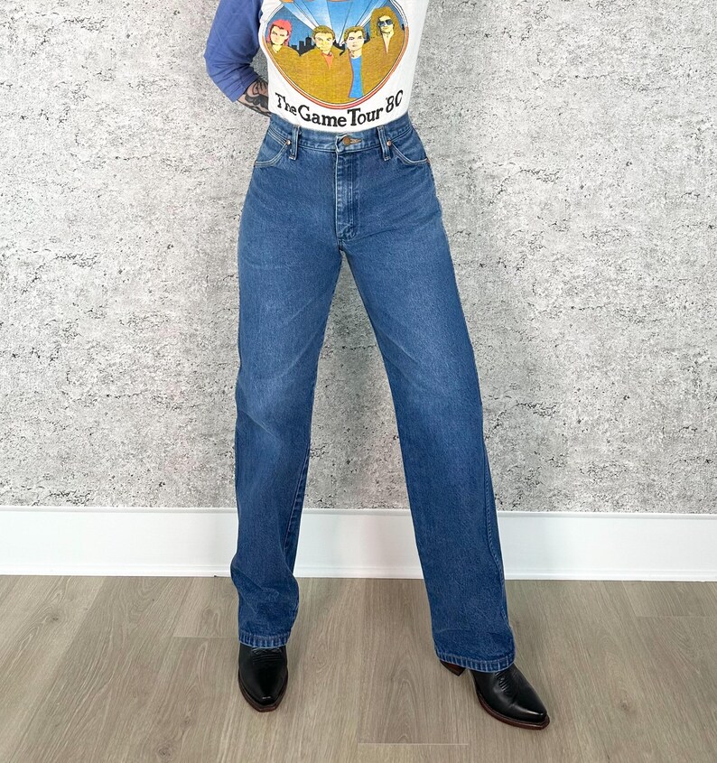 K&ouml;nnte beinhalten: Eine Person tr&auml;gt ein wei&szlig;es und blaues Langarmshirt mit dem Text "The Game Tour 80" und eine blaue Jeans. Die Jeans ist aus mittelblauem Denim mit geradem Bein und wird mit schwarzen Lederstiefeln kombiniert.