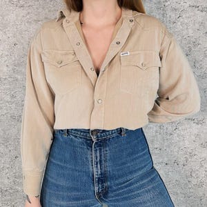 May include: A tan corduroy shirt with snap closures, two chest pockets, and the "GUESS" logo. The shirt is paired with blue denim jeans. The shirt has long sleeves and a collared neckline.