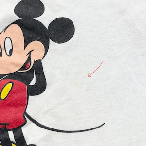 May include: Close-up of a white t-shirt featuring a large graphic of Mickey Mouse. The cartoon character is depicted with black ears, a smiling face, a red jacket, and yellow gloves. The shirt is made of a soft, white fabric.
