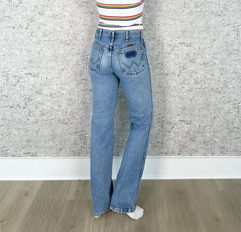Puede incluir: Vaqueros de mezclilla azul claro con pernera acampanada. Los vaqueros tienen un dise&ntilde;o cl&aacute;sico de cinco bolsillos, un parche azul oscuro en el bolsillo trasero y un logotipo de Wrangler. El modelo lleva una camiseta a rayas.