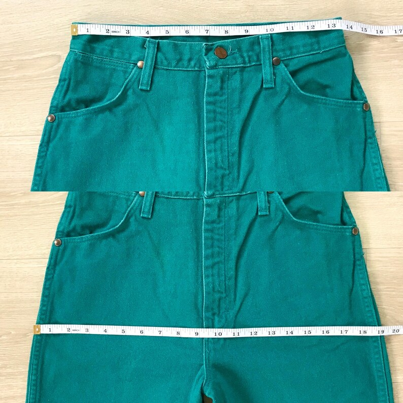 May include: Teal-colored denim jeans with a classic five-pocket design. The jeans are measured with a tape measure, showing a waist measurement of approximately 13 inches. The jeans have a button and zipper closure.
