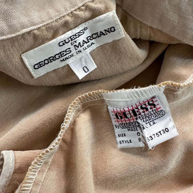 May include: Close-up of two tags from a beige garment. The top tag reads "GUESS by GEORGES MARCIANO MADE IN U.S.A." with a size "0" tag below. The bottom tag reads "GUESS PRODUCTS by GEORGE MARCIANO" with size "0" and style "8 6375T30".