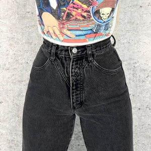 Pu&ograve; includere: Jeans neri a vita alta con chiusura a bottone e tasche anteriori. I jeans sono abbinati a una maglietta grafica bianca con un'illustrazione colorata. I jeans hanno uno stile classico e vintage.