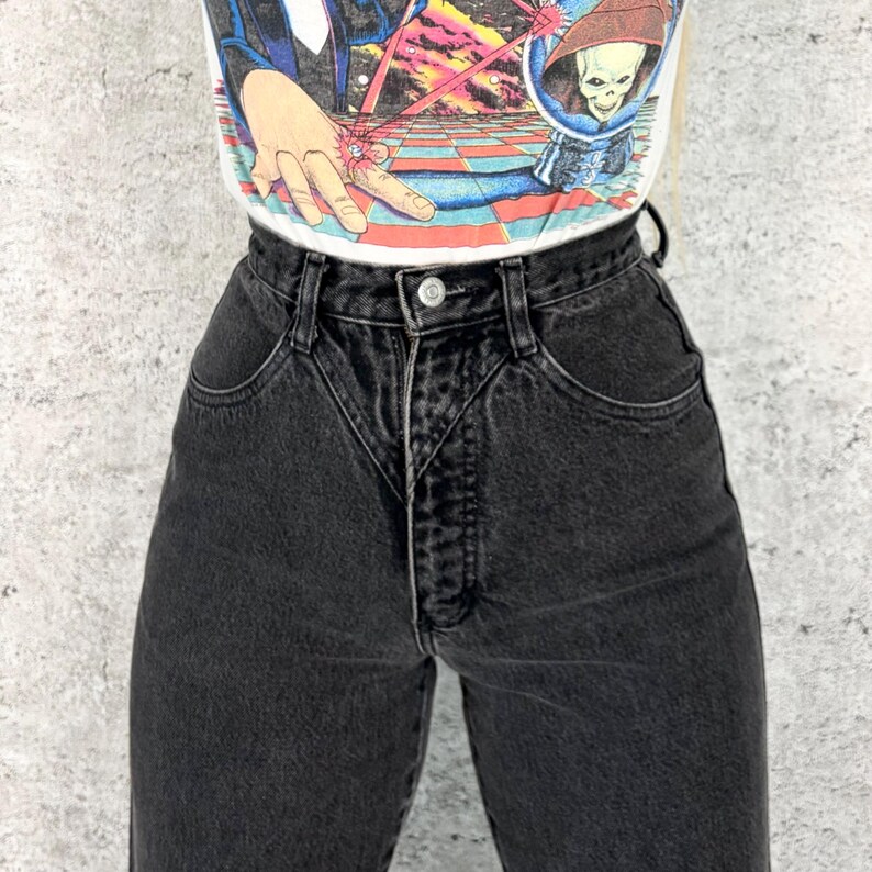 Pu&ograve; includere: Jeans neri a vita alta con chiusura a bottone e tasche anteriori. I jeans sono abbinati a una maglietta grafica bianca con un'illustrazione colorata. I jeans hanno uno stile classico e vintage.