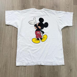 Op de afbeelding: Wit t-shirt met een Mickey Mouse cartoonafbeelding. Het personage wordt getoond in een klassieke pose, met zwarte, rode en gele details. Het shirt is gemaakt van een zacht materiaal, waarschijnlijk katoen.