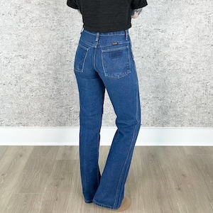 May include: A pair of blue denim jeans with a high waist and a bootcut leg. The jeans have a classic five-pocket design and a small leather patch on the back waistband.