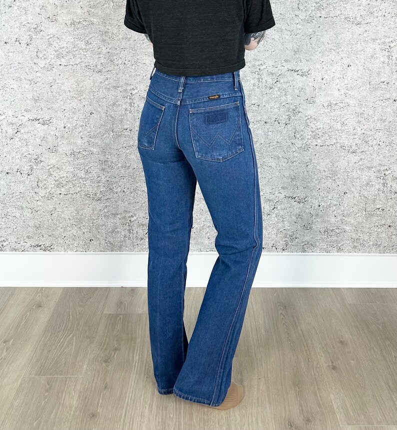 May include: A pair of blue denim jeans with a high waist and a bootcut leg. The jeans have a classic five-pocket design and a small leather patch on the back waistband.