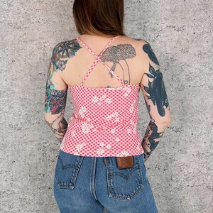 May include: A pink and white gingham print tank top with white daisy floral pattern. The top has a criss-cross back design and thin straps. The model is wearing blue denim jeans.