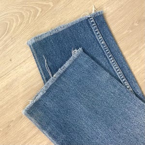 May include: Close-up of blue denim jeans with frayed edges. The jeans are folded, showing the texture and colour of the denim fabric. The frayed edges give the garment a casual, worn appearance.