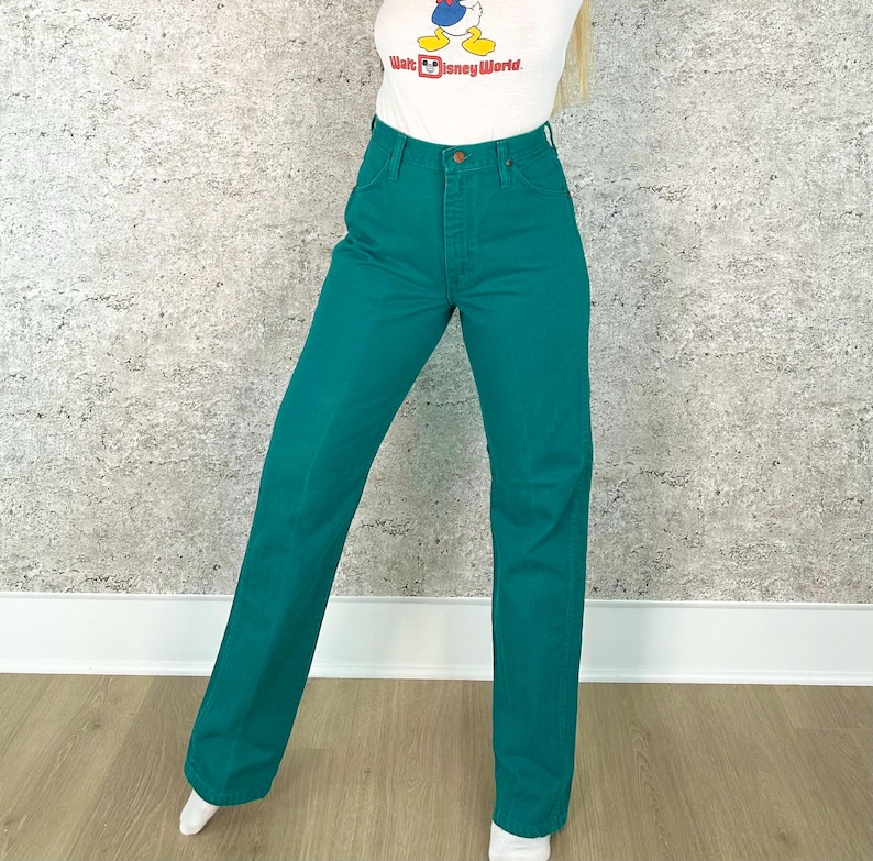 May include: Teal-colored high-waisted jeans. The pants have a classic five-pocket design and a straight leg fit. The jeans are made of a sturdy denim material and feature a button and zipper closure. The model is wearing a white t-shirt with a Walt Disney World graphic.