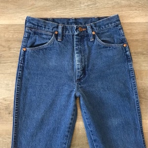 K&ouml;nnte beinhalten: Eine blaue Jeanshose mit hoher Taille und geradem Bein. Die Jeans hat einen klassischen F&uuml;nftaschen-Stil und einen Knopfverschluss aus Kupfer.