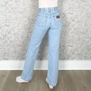 May include: Pale blue denim jeans with a classic fit. The jeans feature a high waist, a brown leather patch on the back pocket, and the Wrangler logo. The jeans have a straight leg and are worn with a white top.