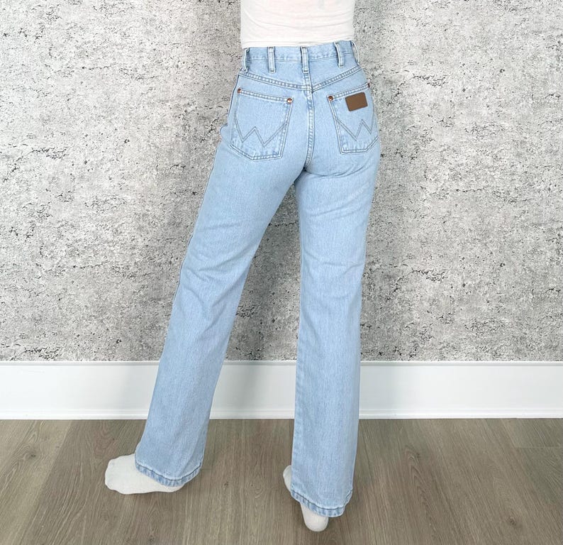 May include: Pale blue denim jeans with a classic fit. The jeans feature a high waist, a brown leather patch on the back pocket, and the Wrangler logo. The jeans have a straight leg and are worn with a white top.