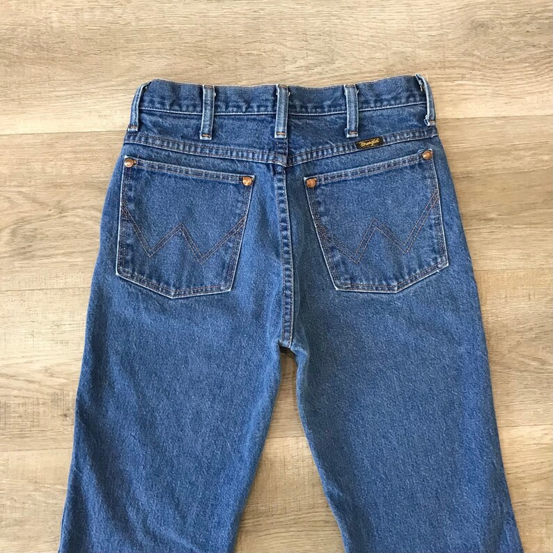K&ouml;nnte beinhalten: Eine blaue Jeanshose mit klassischer gerader Passform. Die Jeans hat einen braunen Leder-Patch mit dem Wrangler-Logo auf dem hinteren Bund. Die Ges&auml;&szlig;taschen haben eine charakteristische W-f&ouml;rmige Steppnaht.