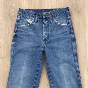 May include: A pair of medium-wash blue denim jeans featuring a high-waisted design, a button fly closure, and copper-toned rivets. The front pockets display light fraying along the edges, and the fabric shows a classic, worn-in texture.