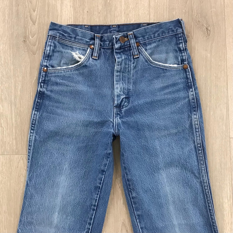 May include: A pair of medium-wash blue denim jeans featuring a high-waisted design, a button fly closure, and copper-toned rivets. The front pockets display light fraying along the edges, and the fabric shows a classic, worn-in texture.