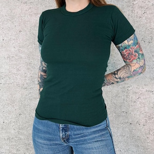 May include: A dark green, short-sleeved t-shirt with a crew neck. The shirt is form-fitting and paired with blue jeans. The sleeves are cuffed. The background is a light grey, textured wall.