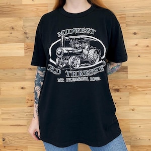 Può includere: Una t-shirt nera a maniche corte con una grafica bianca di un trattore a vapore. Il testo recita MIDWEST OLD THRESHERS MT. PLEASANT, IOWA. Il design è racchiuso in un contorno ovale bianco centrato sulla parte anteriore del capo.