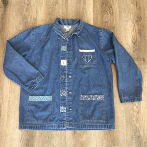 May include: A blue denim jacket with a collared neckline and long sleeves. The jacket features decorative patches, floral pocket accents, and a white pocket with a stitched heart design. It has a button-up front.