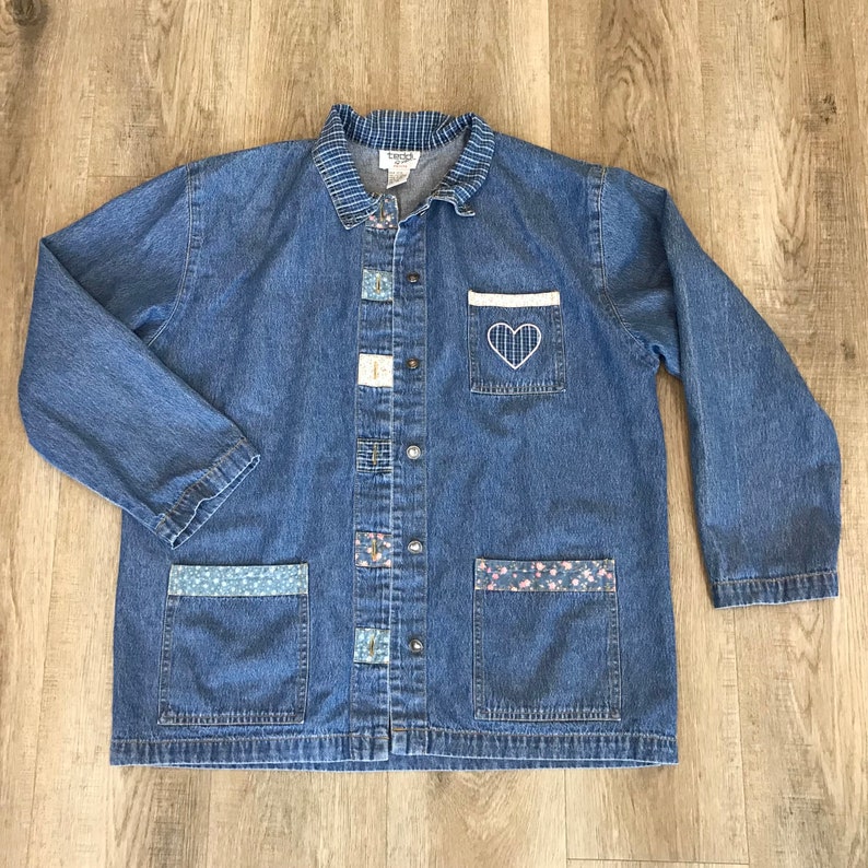 May include: A blue denim jacket with a collared neckline and long sleeves. The jacket features decorative patches, floral pocket accents, and a white pocket with a stitched heart design. It has a button-up front.